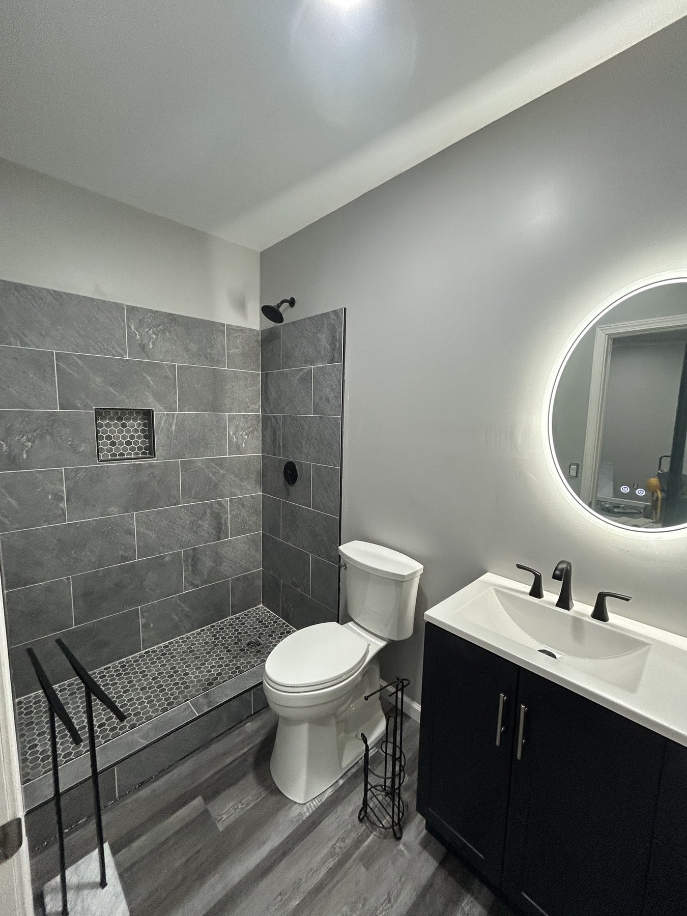 Finished master bath with dark-gray tile walk-in shower, backlit round mirror, and dark vanity