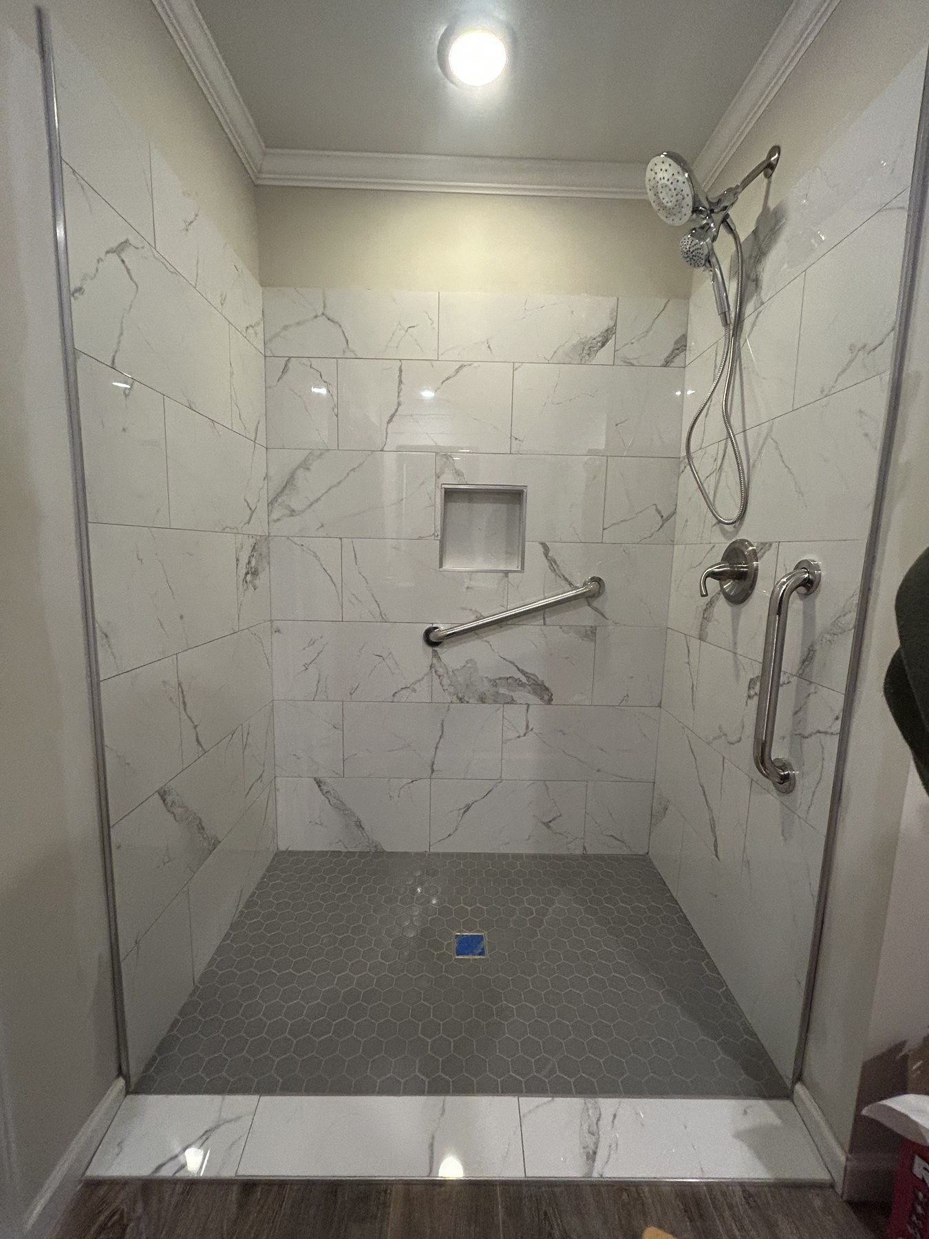 Finished white-tile master walk-in shower with handheld wand, recessed niche, grab bar, and hexagonal mosaic floor