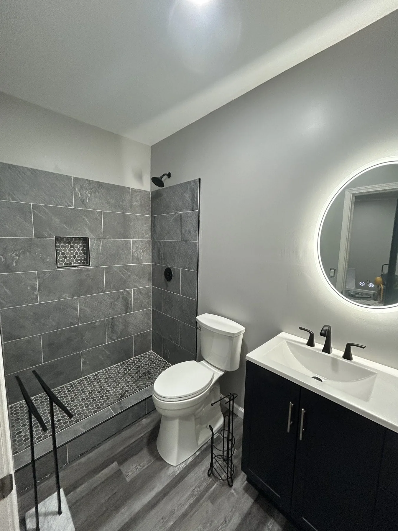 Finished master bath with dark-gray tile walk-in shower, backlit round mirror, and dark vanity